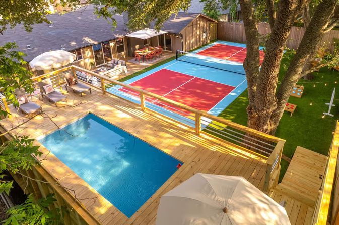 Aerial view of a sunny backyard with a rectangular plunge pool on a wooden deck, lounge chairs and umbrella, and bright red-and-blue pickleball courts under a large shade tree and string lights.