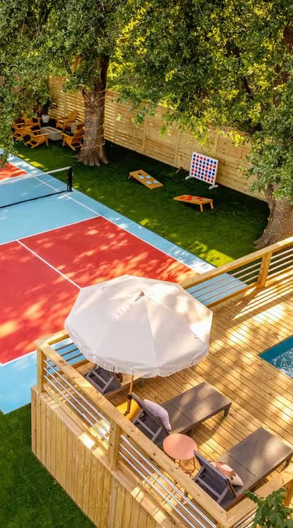 Aerial view of a sunny backyard outdoor-living space with a red-and-blue pickleball/tennis court, wooden deck with a small plunge pool and lounge chairs under an umbrella, dining and lounge seating, lawn games and large shade trees.