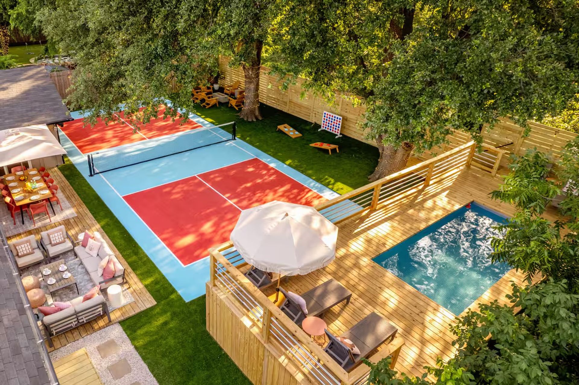 Aerial view of a sunny backyard outdoor-living space with a red-and-blue pickleball/tennis court, wooden deck with a small plunge pool and lounge chairs under an umbrella, dining and lounge seating, lawn games and large shade trees.
