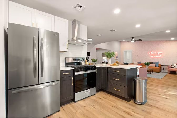 Open-concept modern kitchen with stainless-steel French-door fridge, gas range and hood, dark cabinets with brass pulls, white island countertop and wood floors opening to a cozy living area with a cheery pink accent wall and neon sign.