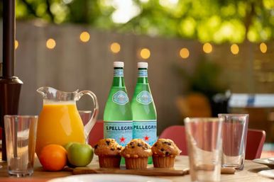 Al fresco patio breakfast table with a pitcher of orange juice, two green sparkling-water bottles, three blueberry muffins on a wooden board, an orange and a green apple, clear drinking glasses and warm bokeh string lights in a blurred backyard.