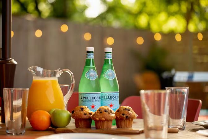 Al fresco patio breakfast table with a pitcher of orange juice, two green sparkling-water bottles, three blueberry muffins on a wooden board, an orange and a green apple, clear drinking glasses and warm bokeh string lights in a blurred backyard.