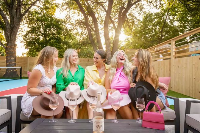 Five friends laughing on a backyard patio at sunset, seated on an outdoor couch with wide‑brim hats, rosé wine glasses and bottle on the table, string lights and trees in the background.