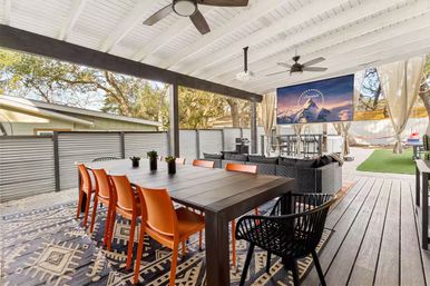 Covered backyard patio with long dark dining table and bright orange chairs, black wicker lounge seating, ceiling fans, mounted projector screen and patterned rug