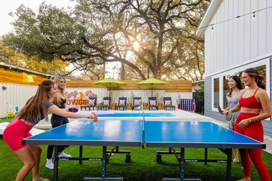 Four friends playing table tennis on a blue ping-pong table at a poolside backyard patio with artificial turf, lounge chairs, green umbrellas, string lights and oak trees glowing at golden hour