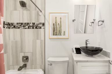 Bright modern bathroom with wood-look tile shower and mosaic accent, white toilet, marble vanity with black glass vessel sink and playful cactus wall art.