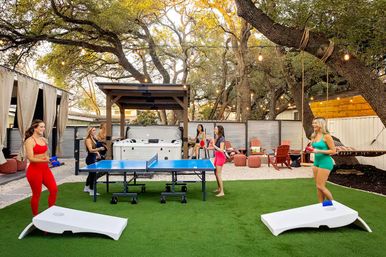 Women in colorful activewear playing ping-pong and cornhole on artificial turf in a backyard entertainment space with a hot tub, hammock, string lights, and patio seating under oak trees.