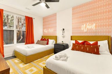 Cheerful modern guest room with two mustard-yellow platform beds, coral-patterned accent walls with neon "Weird" sign, orange curtains, geometric rug, and black nightstand