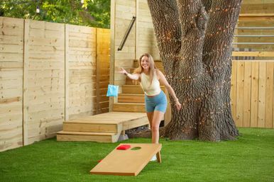Smiling woman tossing a blue bean bag at a cornhole board on artificial turf in a fenced backyard, beside a large tree wrapped in string lights and wooden deck steps.