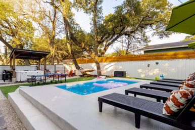 Sunlit backyard with rectangular plunge pool and pink float, row of black lounge chairs with patterned pillows on a concrete deck, pergola with outdoor kitchen and hot tub, ping-pong table and hammock under mature trees