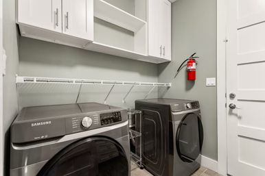 Modern residential laundry room with matching Samsung dark-gray front-load washer and dryer, white wall cabinets and wire shelf, mounted red fire extinguisher, pale green walls and white door.