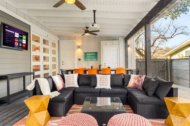 Cozy covered Austin backyard patio with black wicker sectional and 'HOWDY' throw pillows, glass coffee table, orange dining chairs, wall-mounted TV, ceiling fans and Austin city limit sign.
