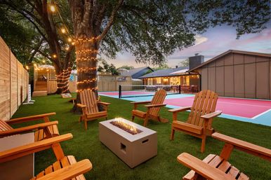 Evening suburban backyard entertaining space with Adirondack chairs around a modern rectangular fire pit, string lights on a large tree, and a colorful pink-and-blue pickleball court next to a lit patio.