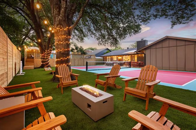 Evening suburban backyard entertaining space with Adirondack chairs around a modern rectangular fire pit, string lights on a large tree, and a colorful pink-and-blue pickleball court next to a lit patio.