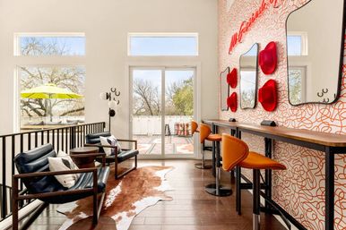 Sunlit modern lounge with sliding glass door to a balcony, navy leather chairs, cowhide rug, lime-green patio umbrella outside, orange bar stools and playful red hat mirrors on a patterned accent wall.