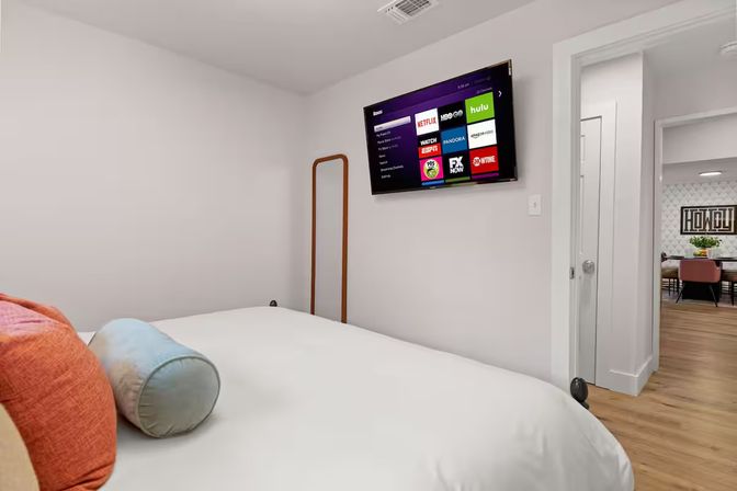Cozy modern bedroom with white bedding, colorful throw pillows, wall-mounted smart TV displaying streaming apps, full-length mirror and open doorway to a wood-floored dining area.