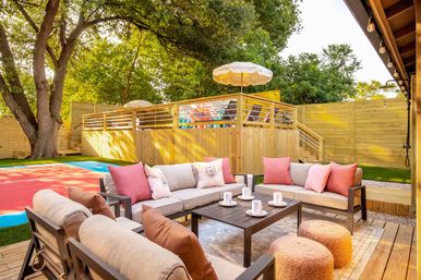 Cozy sunny backyard patio with cushioned sofas and pink throw pillows around a central coffee table, woven poufs, wooden deck, raised deck with umbrella, privacy fence and large shade trees