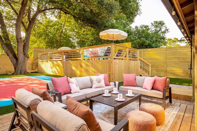 Cozy sunny backyard patio with cushioned sofas and pink throw pillows around a central coffee table, woven poufs, wooden deck, raised deck with umbrella, privacy fence and large shade trees