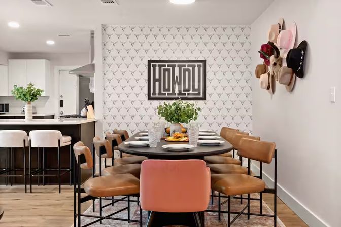 Open-concept dining room and kitchen with round black table set for eight, tan leather chairs and a pink accent chair, Howdy wall art over longhorn-patterned wallpaper, cowboy hats on the wall, kitchen island with white bar stools and hardwood floors