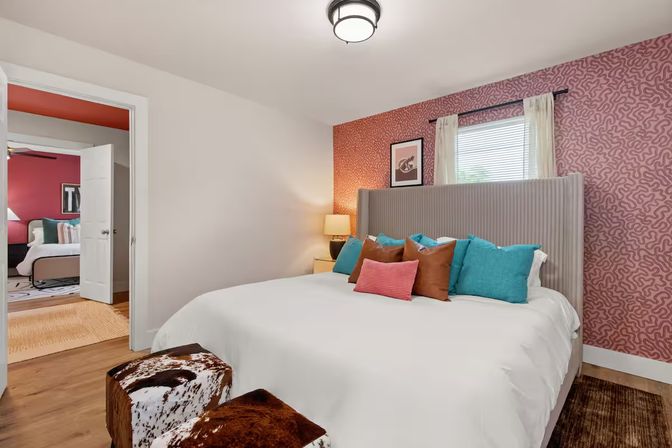 Bright contemporary bedroom with white bedding, tall gray upholstered headboard, pink patterned accent wall, teal and rust throw pillows, cowhide ottomans on wood floor, bedside lamp and open door to an adjoining room