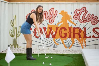 Smiling woman in denim skirt and cowboy boots putting on an outdoor mini-golf green in front of a colorful "Cowgirls" mural featuring a horse silhouette and cactus