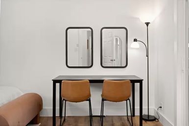 Minimalist apartment workspace with a black desk and two tan leather chairs, twin black-framed mirrors on a white wall, arched floor lamp and warm hardwood floor.