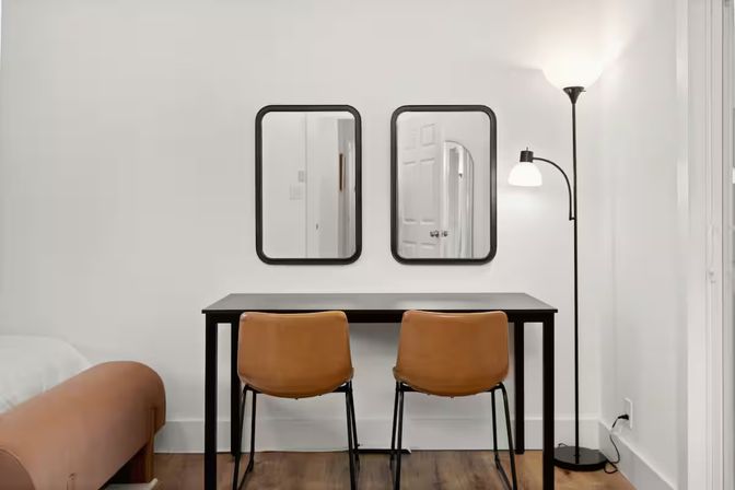 Minimalist apartment workspace with a black desk and two tan leather chairs, twin black-framed mirrors on a white wall, arched floor lamp and warm hardwood floor.
