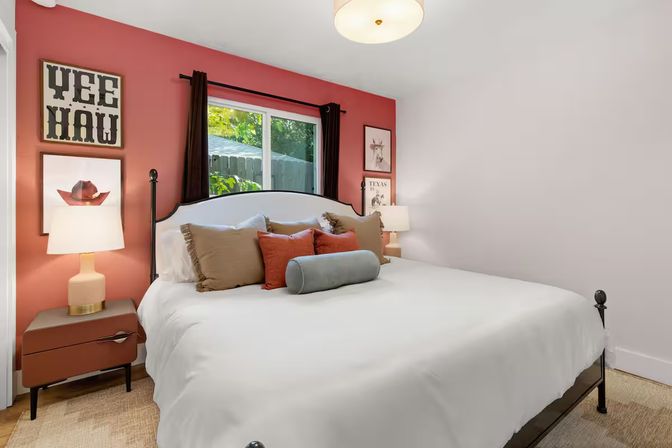 Cozy Texan-inspired bedroom with coral-red accent wall, black metal bed frame, crisp white bedding, layered throw pillows, matching nightstands and lamps, and a window overlooking a backyard.