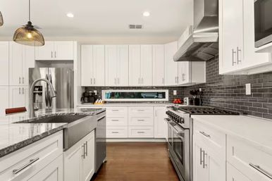 Bright modern open-concept white kitchen with large island and farmhouse sink, stainless steel refrigerator and range, gray subway tile backsplash, pendant lighting and warm hardwood floors.