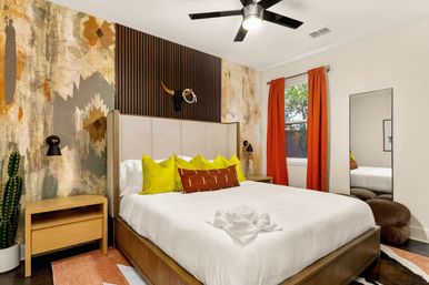 Modern boho bedroom with wood platform bed, textured Southwestern-style accent wall, bright lime-yellow throw pillows, rust-orange curtains, black ceiling fan, potted cactus and full-length mirror.