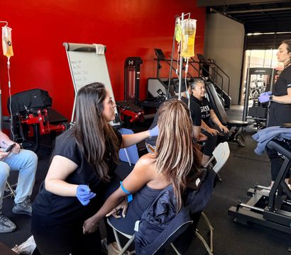 Women receiving IV vitamin drips at a pop-up wellness station inside a gym, clinicians in gloves attending clients against a bright red wall with exercise equipment in the background.