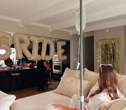 Person reclining on a beige sofa with an IV drip pole nearby, gold inflatable "BRIDE" balloons hanging above a kitchen in a cozy living-room bridal celebration.