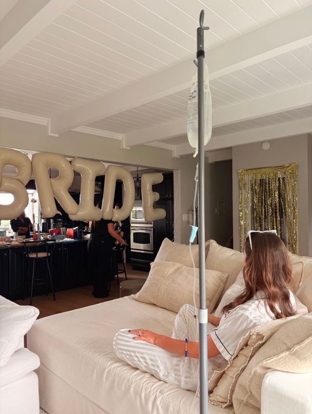 Person reclining on a beige sofa with an IV drip pole nearby, gold inflatable "BRIDE" balloons hanging above a kitchen in a cozy living-room bridal celebration.