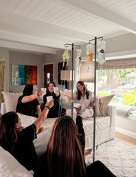Group of friends on a white sectional in a sunlit living room toasting cups while receiving IV hydration drips on poles — fun at-home wellness IV drip party.