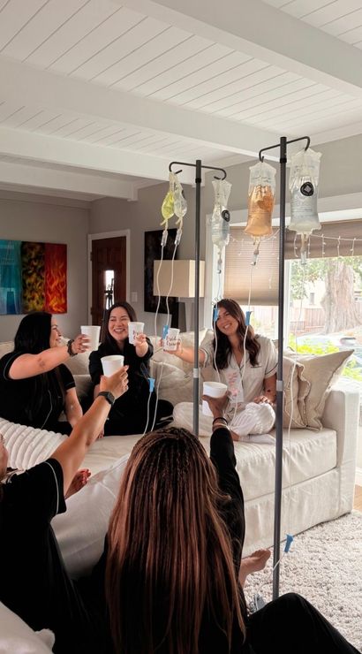 Group of friends on a white sectional in a sunlit living room toasting cups while receiving IV hydration drips on poles — fun at-home wellness IV drip party.