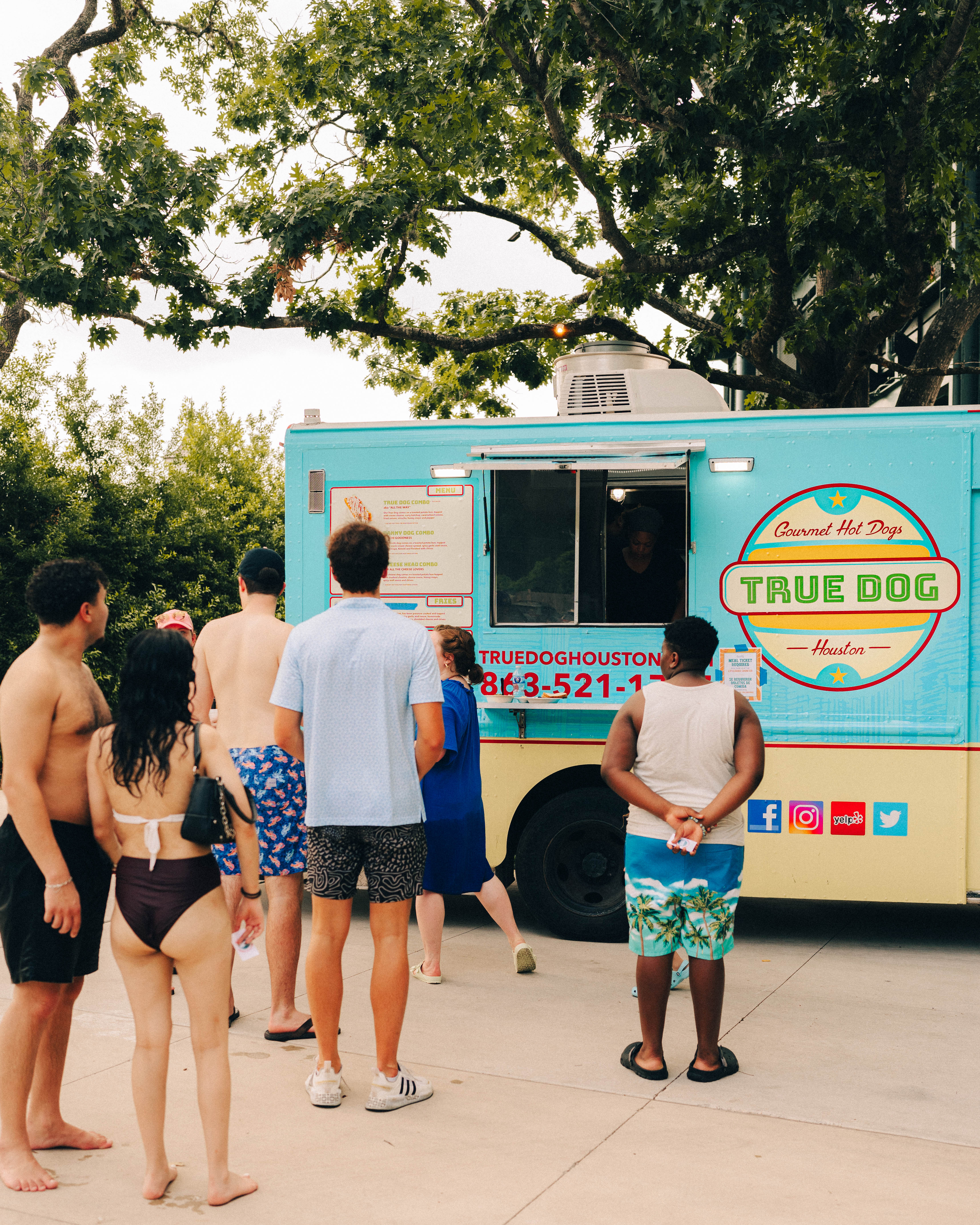 Summer scene at a turquoise food truck serving gourmet hot dogs in Houston, with people in swimsuits and summer clothes queuing beneath shady trees.