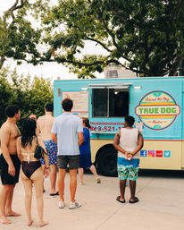 Summer scene at a turquoise food truck serving gourmet hot dogs in Houston, with people in swimsuits and summer clothes queuing beneath shady trees.