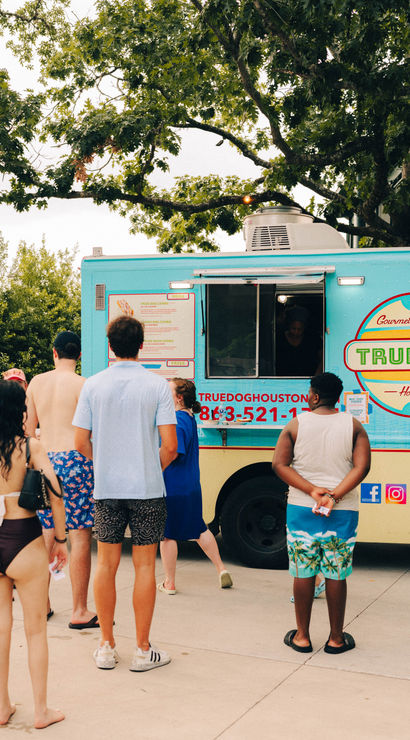 Summer scene at a turquoise food truck serving gourmet hot dogs in Houston, with people in swimsuits and summer clothes queuing beneath shady trees.