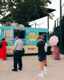 Bright turquoise food truck serving gourmet hot dogs at an outdoor stadium entrance, with a small crowd queuing—a teen in sneakers, a man in uniform, and a woman holding a pink umbrella under nearby trees.