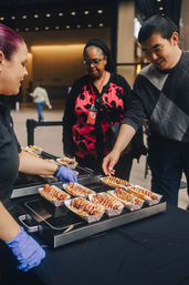 Customers at an indoor food stall selecting rows of sauce‑drizzled gourmet hot dogs in paper trays, vendor wearing gloves serving sausages from a heated tray — indoor market/street food scene.
