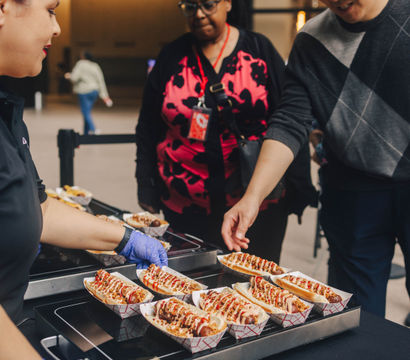 Customers at an indoor food stall selecting rows of sauce‑drizzled gourmet hot dogs in paper trays, vendor wearing gloves serving sausages from a heated tray — indoor market/street food scene.