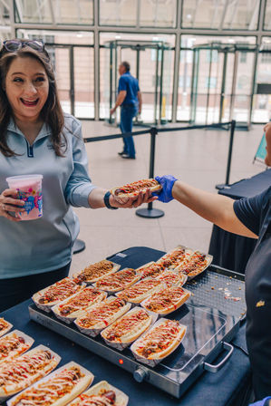 Smiling attendee at an indoor urban atrium accepting a loaded gourmet hot dog from a gloved vendor, tray of topped hot dogs on the table and a cup of cotton candy in hand.