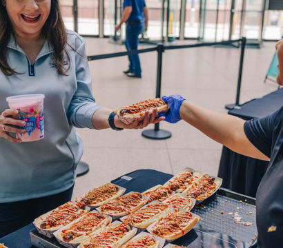Smiling attendee at an indoor urban atrium accepting a loaded gourmet hot dog from a gloved vendor, tray of topped hot dogs on the table and a cup of cotton candy in hand.