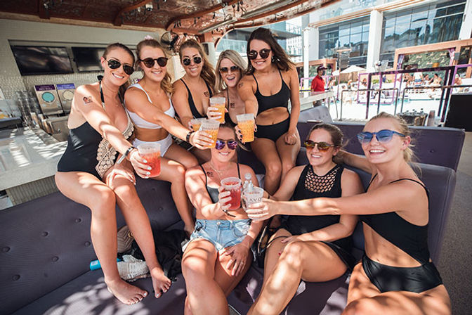 Eight women in swimsuits and sunglasses toasting with colorful cocktails in a sunny poolside cabana lounge at a resort pool.