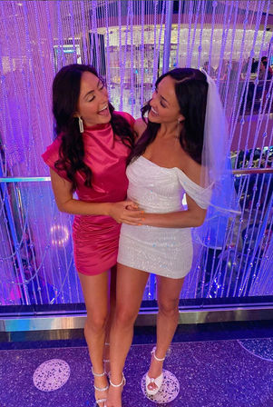 Two women laughing at a bachelorette party — one in a white sequined mini dress and veil, the other in a hot-pink satin mini dress, posing arm-in-arm in front of a sparkling beaded curtain with purple club lighting.