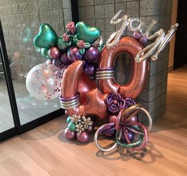 Metallic rose-gold '40' balloon display with silver 'yay' script, teal heart balloons, confetti globe and colorful floral clusters staged in an indoor event lobby on a wood floor.