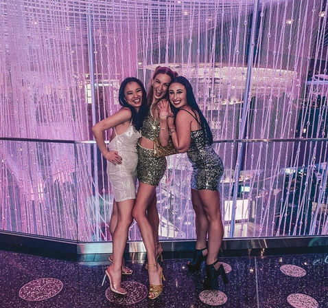 Three women in sequined party dresses and heels laughing and posing on a balcony against a purple-pink curtain of hanging crystal beads and lights in a glamorous nightclub-like venue.