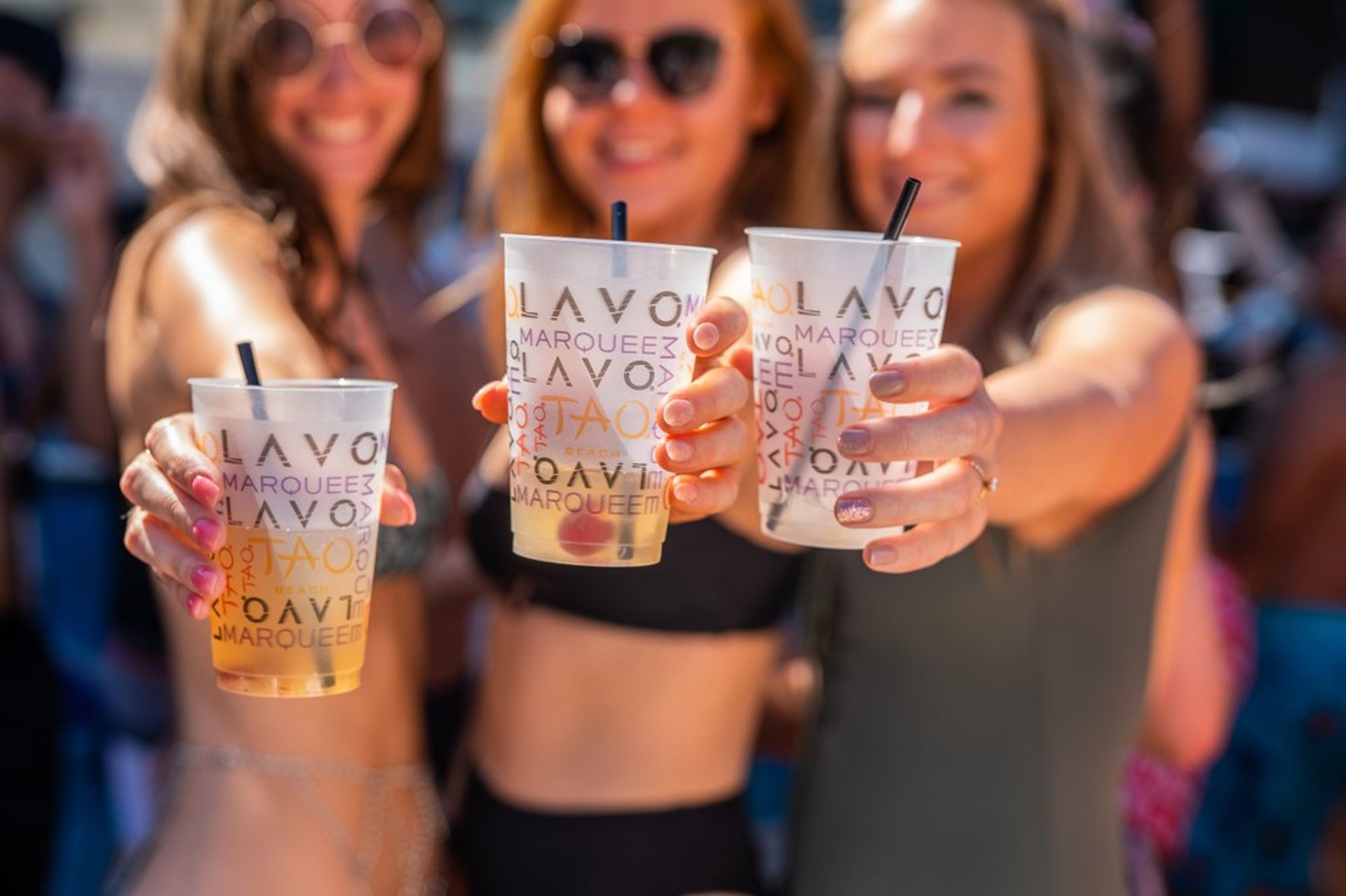 Three women in swimsuits and sunglasses toasting with clear plastic drink cups toward the camera at a sunny outdoor pool party.