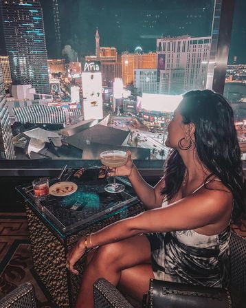Woman sipping a cocktail at a rooftop lounge overlooking the neon-lit Las Vegas Strip skyline at night, sparkling city lights and busy streets below.