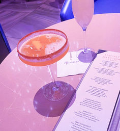 Two cocktails on a marble table in a neon-pink upscale lounge: a salt-rimmed citrus coupe and a champagne flute beside a cocktail menu